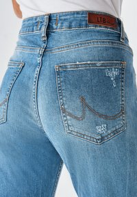 Gaišzilā džinsu bikses ar vidēju vidukļa augstumu, kontrastējošām oranžām šuvēm un divām aizmugurējām kabatām ar izteiktu dizainu.