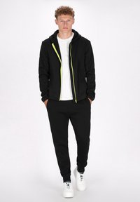 Zwarte zip-up hoodie met een limoen groene rits, gecombineerd met bijpassende zwarte joggers. Gelaagd over een wit shirt, gedragen met witte sneakers.