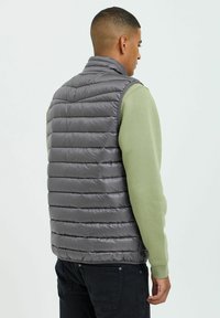 Chaleco acolchado gris con textura de rombos horizontales, cuello alto, usado sobre una camisa de manga larga verde claro, presenta una silueta ajustada.