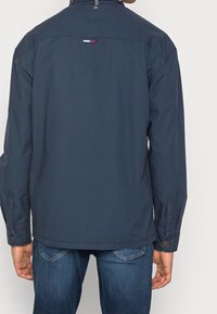 Mörkblå jacka i ett slätt material med krage, synliga stygn och knapprenar på ärmarna. Bakom finns en liten färgblockdetalj.