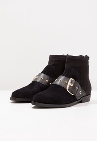 Botas de tobillo de ante negro con una correa negra mate que presenta herrajes dorados. Punta redondeada y tacón bajo. Parte superior texturizada con detalles suaves.