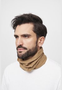 Hombre con barba y cabello oscuro, llevando un pasamontañas beige y camisa blanca, mirando con concentración frente a un fondo liso y claro.