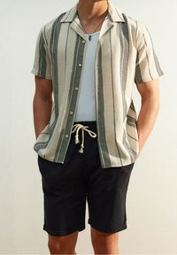 Man in een beige overhemd met korte mouwen en verticale strepen, een witte tanktop, zwarte shorts met trekkoord en een hangerketting.