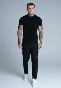 Camiseta de algodón negra con mangas cortas, combinada con pantalones cargo negros con bolsillos laterales y zapatillas blancas, sobre un fondo gris claro.