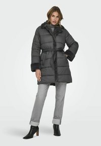 Abrigo gris acolchado con gran cuello y ribete de piel sintética, ceñido en la cintura, combinado con jeans gris claro con vuelta en los puños y botas de tacón negras.