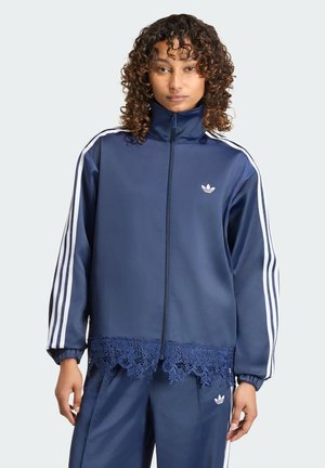 Žena nosí tmavě modrou teplákovou soupravu Adidas s bílými pruhy na rukávech a kalhotách a krajkovým lemem na spodním okraji bundy.