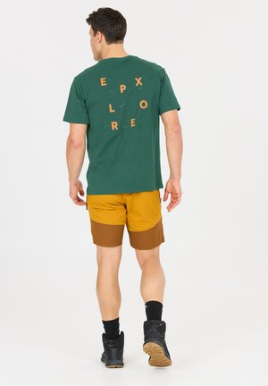 Mann, der weggeht, trägt ein grünes T-Shirt mit dem Schriftzug „EXPLORE“ und senfgelbe Wandershorts, schwarze Socken und schwarze Stiefel.
