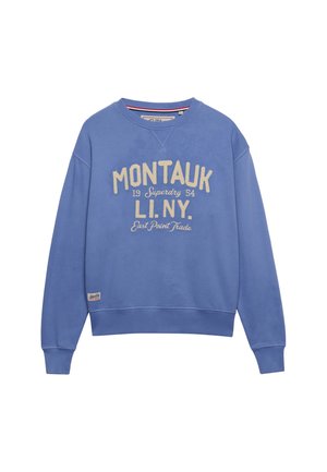 Blå crewneck sweatshirt med beige tekst "MONTALIK 19 Superdry 54 LI. NY. East Point Trade" og en lille brandpatch nær kanten.