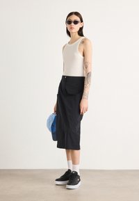 Gonna cargo nera con tasche, abbinata a una canotta bianca. Gli accessori includono un cappellino azzurro chiaro e sneakers nere. Gli occhiali da sole completano il look.