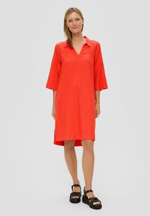 Rotes Leinenkleid mit Dreiviertelärmeln, V-Ausschnitt und Seitenschlitzen. Besitzt eine lockere Passform und eine glatte Textur. Wird mit schwarzen Plateausandalen getragen.
