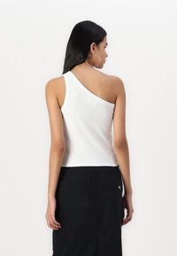 Débardeur blanc à côtes avec une seule bretelle, associé à une jupe noire comportant deux poches arrière et un détail de logo. Texture lisse.