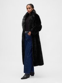Manteau en fausse fourrure noire, design long avec grand col et manches surdimensionnées, associé à un jean bleu taille haute et des bottines noires.