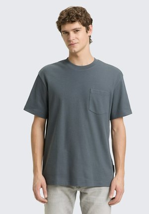 Jonge man draagt een eenvoudige grijze T-shirt met korte mouwen en een borstzakje, gecombineerd met lichtgrijze broek, staand tegen een neutrale achtergrond.