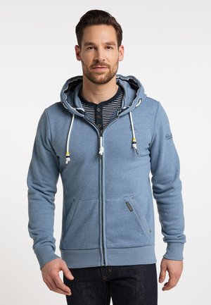 Mann trägt hellblauen Kapuzenpullover mit Reißverschluss und weißen Kordeln über einem schwarz-weiß gestreiften Henley-Shirt und steht vor weißem Hintergrund.