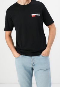 Camiseta de algodón negra con un pequeño logo en rojo, blanco y negro en el lado izquierdo del pecho. Mangas cortas, cuello redondo, corte relajado.