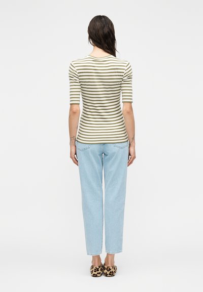 Άτομο με μακριά σκούρα μαλλιά, ντυμένο με κοντομάνικο μπλουζάκι με πράσινες και λευκές ρίγες, ανοιχτόχρωμο τζιν και παντόφλες με εκτύπωση λεοπάρδαλης, στέκεται με την πλάτη προς τα εμάς.