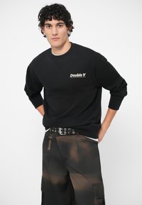 Μαύρο φούτερ με στρογγυλή λαιμόκοψη και ριμπ μανσέτες, με μικρό λευκό λογότυπο. Συνδυάζεται με σκούρα cargo παντελόνια με σχέδιο tie-dye.