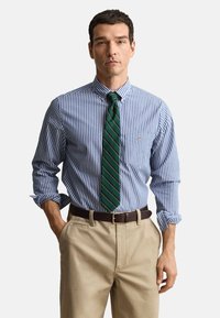 Koszula w niebiesko-białe paski z kołnierzykiem zapinanym na guziki, zestawiona z zielono-złotym krawatem w paski. Beżowe spodnie i brązowy pasek.