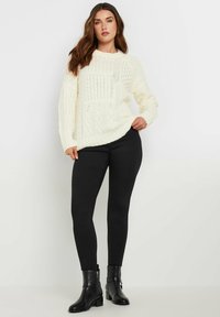 Crème gebreide trui met een gestructureerd kabelpatroon, ronde halslijn en ribbelmanchetten, gecombineerd met zwarte leggings en enkellaarsjes.