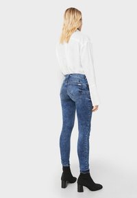 Vaqueros skinny de mezclilla azul de talle alto con un acabado desteñido, combinados con botas negras hasta el tobillo, vistos desde atrás contra un fondo neutro.