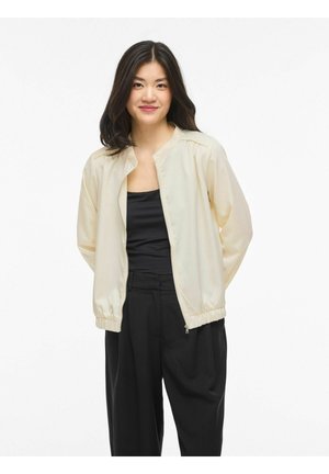 Vrouw met lang donker haar, gekleed in een crème kleurige jas met rits over een zwarte top en zwarte wijde broekspijpen, staande tegen een witte achtergrond.