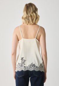 Camisole couleur crème avec des bretelles spaghetti, présentant une finition en dentelle à l'ourlet. Tissu doux et coupe décontractée, associée à un jeans bleu foncé.