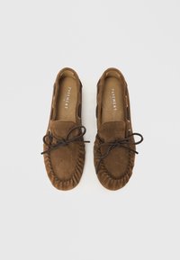 Mocassins en daim marron avec détail de lacets, bout arrondi, surpiqûres décoratives et accents découpés sur les côtés. Semelles plates et texture souple.