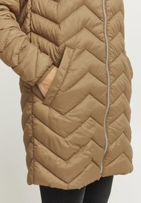 Brun chevron-quiltet pufferjakke med lynlås, som har sidelommer og en glat, let skinnende tekstur.