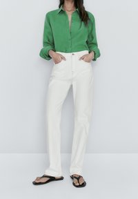Woman standing with hands in pockets wearing green button-up shirt, white high-waisted jeans, and black thong sandals against a plain wall.