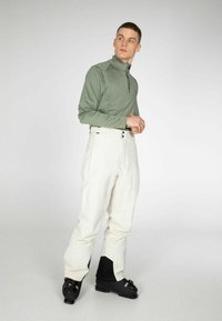 Groene lange mouw zip-up top gecombineerd met witte sneeuwbroeken en zwarte skischoenen. De top heeft een hoge kraag en platte naden voor comfort.