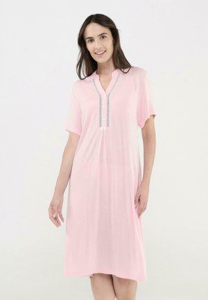 Mujer de pie sonriendo, vestida con un vestido rosa suelto hasta la rodilla, con mangas cortas y botones en la parte delantera.