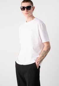 Bílý bavlněný tričko s světle modrým logem na hrudi, spárované s černými kalhotami. Doplňky zahrnujíOversized sluneční brýle a tetování na ruce.