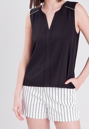 Vrouw draagt een mouwloze zwarte blouse met V-hals en witte shorts met dunne verticale zwarte strepen, één hand in de zak.