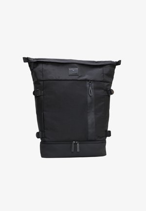 Sac à dos en tissu noir avec un fond roulé, finition lisse, sangles latérales réglables, poche zippée à l'avant et base rectangulaire. Étiquette de la marque sur le devant.