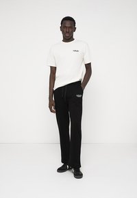 Balta medvilninė marškinėliai su mažu juodu grafiniu paveikslėliu ant krūtinės, derinami su juodomis sportinėmis kelnėmis, kurių priekyje yra logotipas, vilkimi su juodais sportbačiais.