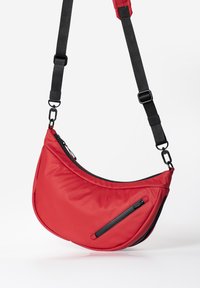 Rote Stoff-Schultertasche mit einer geschwungenen Form, schwarzen Reißverschlussakzenten und verstellbarem schwarzen Tragegurt. Enthält eine vordere Reißverschlusstasche.