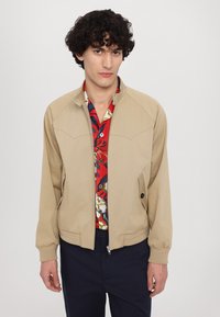 Béžová bomber bunda se zipem, dvěma bočními kapsami, ribbed manžetami a detaily na ramenech, nošená přes červenou květovanou košili.