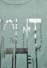 Hellgrünes Baumwoll-T-Shirt mit silbernem Metallic-Text in verschiedenen Schriftarten, einschließlich Phrasen wie "reflektiere und scheine" und "lass deine Seele strahlen."