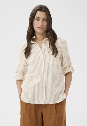 Kobieta z długimi brązowymi włosami, ubrana w beżową pasiastą koszulę na guziki z podwiniętymi rękawami oraz brązowe spodnie, stoi z rękami w kieszeniach.