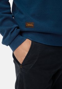 Navy textured sweater with ribbed cuffs, paired with black pants featuring a gold accent button and side pocket detail.