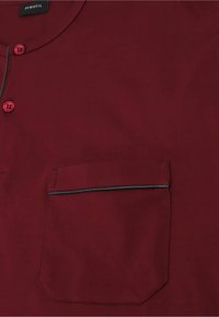 Camicia bordeaux a maniche corte con scollatura rotonda, caratterizzata da due bottoni rossi e una tasca sul petto bordata da un sottile bordo nero.