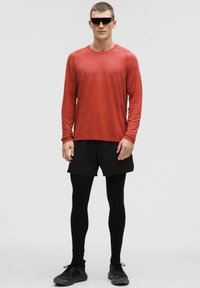 Tricou sport roșu cu mânecă lungă, pantaloni scurți negri, colanți negri și adidași. Modelul poartă ochelari de soare închiși la culoare. Design simplu și funcțional.