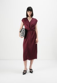 Nainen, jolla on yllään hihaton burgundinpunainen midi-mekko, pitää suurta harmaata käärmeennahkakuosisista clutch-laukkua, ja hänellä on mustat kärjelliset korkokengät.
