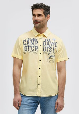 Mann, der ein hellgelbes kurzärmeliges Hemd mit blauem Aufdruck und blaue Jeans trägt, vor einem einfarbigen Hintergrund steht.