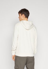 Felpa beige chiaro con cappuccio regolabile, maniche lunghe, polsini e orlo a coste, realizzata in un tessuto morbido e testurizzato, abbinata a pantaloni cargo grigi.