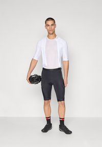 Completo da ciclismo che include una maglia attillata bianca a maniche corte e pantaloni neri imbottiti. Il modello tiene in mano un casco nero, indossando scarpe da ciclismo nere.