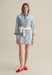 Denim jurk met lange mouwen, twee voorzakken en een knoopsluiting; taille cinched met een witte trui geknoopt om de heupen, gecombineerd met rode ballerina's.