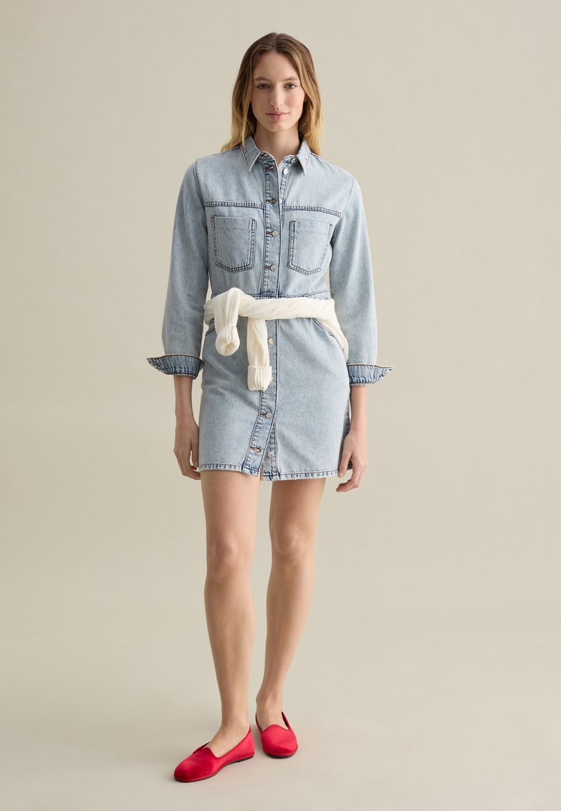 Denim jurk met lange mouwen, twee voorzakken en een knoopsluiting; taille cinched met een witte trui geknoopt om de heupen, gecombineerd met rode ballerina's.