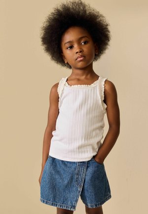 Ung pige med naturligt afro-hår iført en hvid ærmeløs top med blonderkant og denimshorts, stående med den ene hånd i lommen.