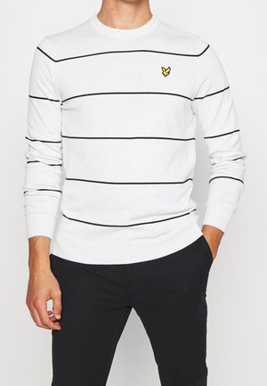 Mand iført en hvid langærmet sweater med tynde sorte vandrette striber og et lille gult fuglelogo på brystet, kombineret med sorte bukser.